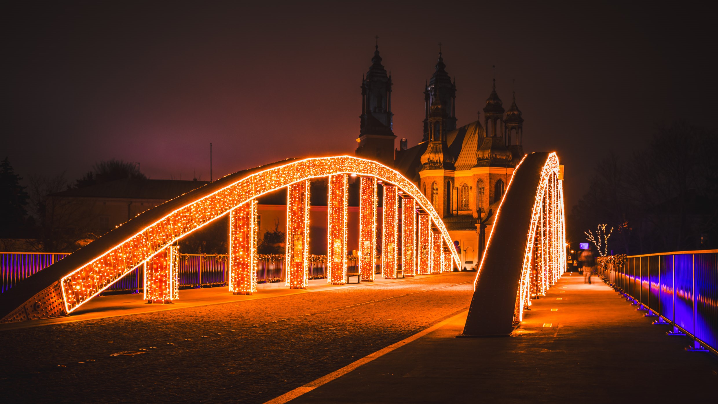 Zdjęcie przedstawia most udekorowany świąteczną iluminacją, z kościołem w tle.